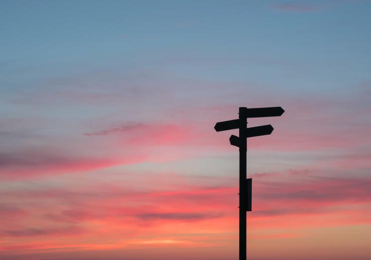 Foto på siluett av skylt med pilar i fyra olika riktningar. I bakgrunden himmel med blå och rosa skymning.